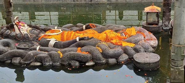 Stone sculpture of Lord Vishnu reclining on the serpent Shesha, partially submerged in a pond, adorned with orange cloth and garlands, at Budhanilkantha Temple in Kathmandu, Nepal.